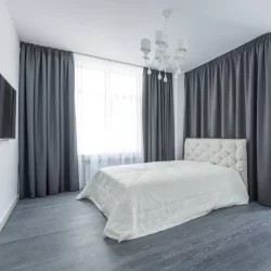 White bed with grey curtains in a bedroom with sheer curtain